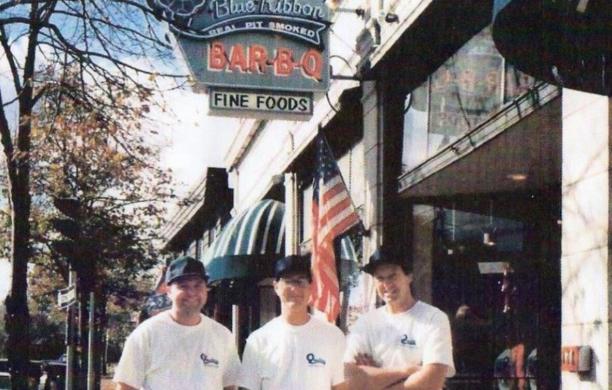 Three people in white shirts and caps stand smiling in front of a barbecue restaurant.