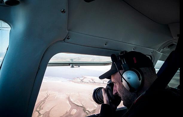 The camera is over the shoulder of a man wearing a baseball hat looking into a professional camera pointed out the window of an airplane in flight over dry land..