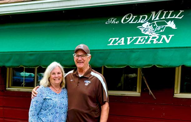 In front of a green awning with “Old Mill Tavern” printed in white, a woman and a man stand with their arms around each other’s shoulders.