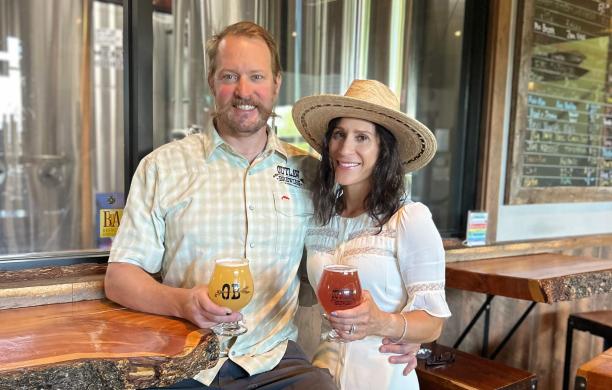 Todd Hough sits on a bar stool at a table made from salvaged wood, while Nicole stands beside him, each holding a glass of beer.