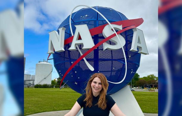 A woman stands smiling in front of a large NASA logo sculpture outdoors on a grassy area with blue sky and clouds.