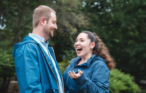 Against a backdrop of lush green foliage, Sara Anderson is facing Christian Hillegas with her mouth smiling a gape in surprise and holding one hand out.