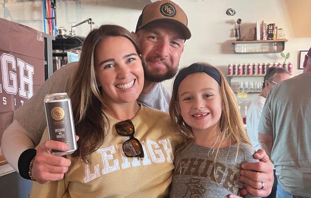 Mike, his wife, and their daughter smile with their arms around each other wearing Lehigh sweatshirts. The man holds a beer can labeled as Brown and White 