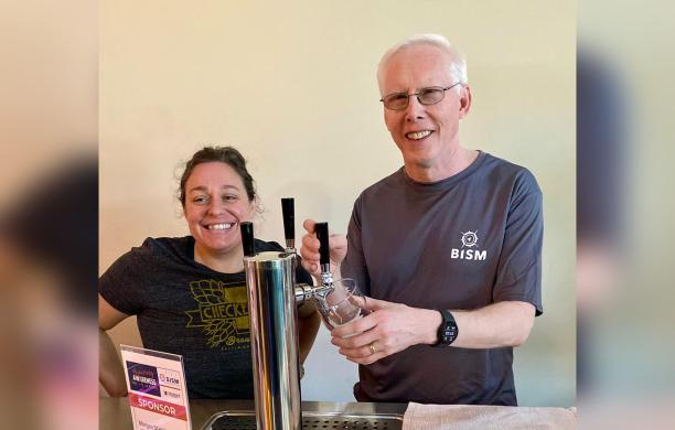 Standing beside a woman behind a beer tap, Mike Gosse pours beer into a pint glass, wearing a dark gray B.I.S.M. t-shirt.
