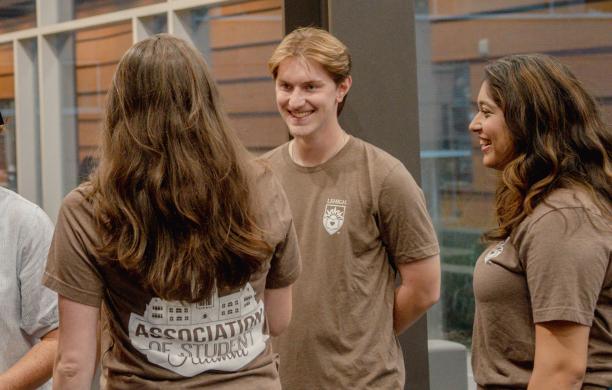 Students wearing brown shirts that read "Association of Student Alumni" socialize in the corridor of a campus building.