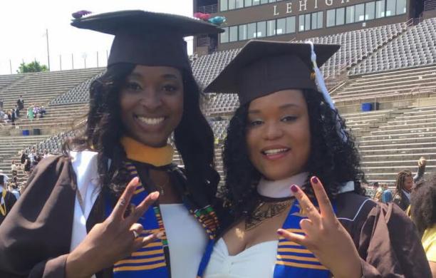 Two smiling women in graduation caps, gowns, and stand closely and show hand signals in relation to their sorority.