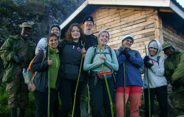 A group of 9 hikers pose, dressed warmly and holding bamboo as hiking sticks in front of a rustic wood cabin.