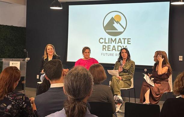 Four women sit on a stage. One holds a microphone. A slide behind them says "Climate Ready"