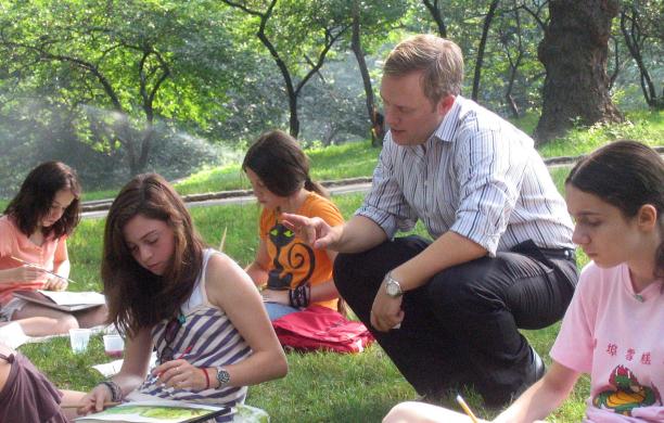 Squatting in the grass next to one female student along a few sitting in the grass, William Crow advises technique with his long sleeves rolled up.