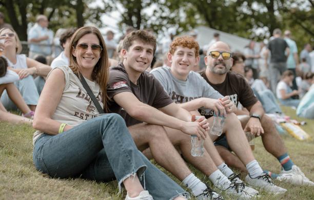 Sitting on a sloped grassy lawn, a family of four smile for a photo with two sons in the center and mom and dad on either side.