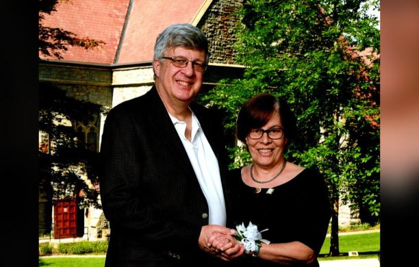 Steve Villas and wife stand holding hands in front of a backdrop showing a stone-faced building.