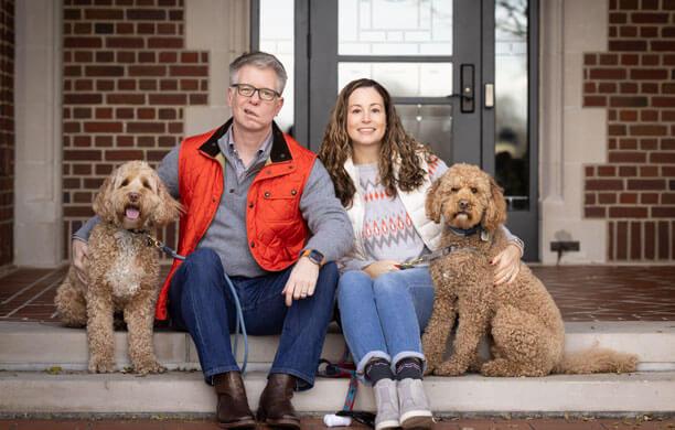 A man and a woman sit on the porch of their home and smile with their two dogs.