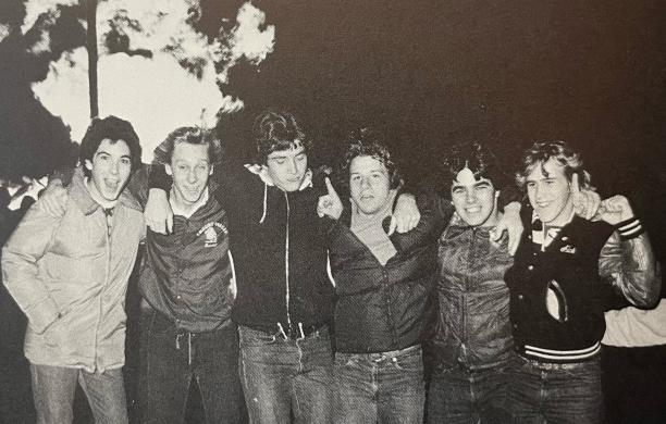 In a black and white photo, six young men in jackets smile arm-in-arm with a large fire shown blazing behind them.
