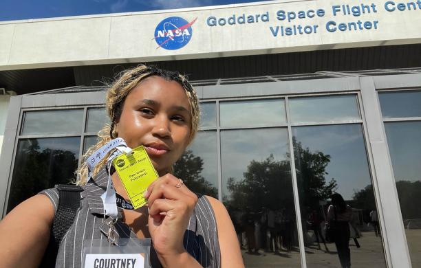 Courtney Baker stands in front of a NASA building and holds a yellow event badge that lists the name of the fellowship. 