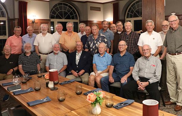 A group of older men sit and stand around a large table