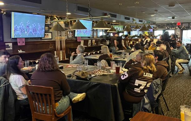 A group of alumni wearing brown and white sit around tables in a restaurant watching the Rivalry game.