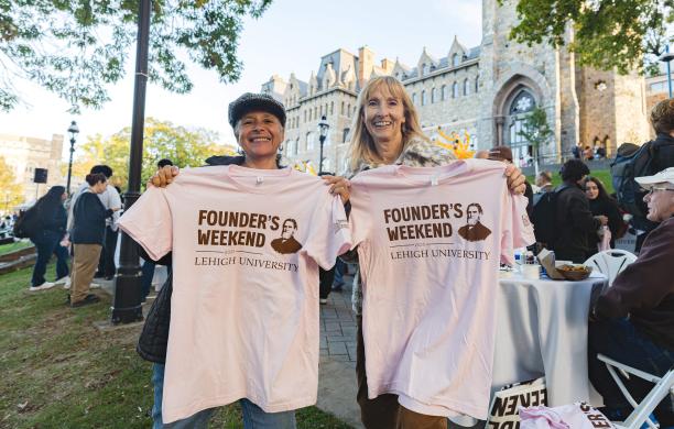 Two alumnae stand on the front lawn of the Clayton UC with the building in the distance holding up baby pink tshirts that read “Founder’s Weekend Lehigh University.”