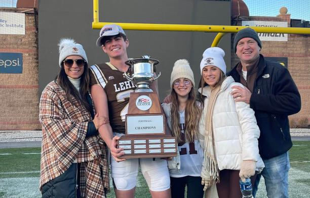A Lehigh football player holding a large championship trophy stands on the field with four smiling family members.