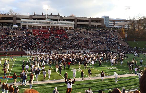 Dozens of fans swarm the field post Lehigh's win, cheering, while other fans fill the stands. Nearby, Clutch cheers.
