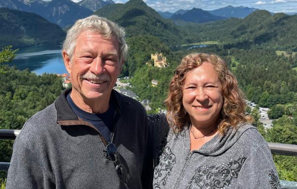 A man and woman pose for a photo on a bridge, smiling with a green, mountainous landscape behind them.