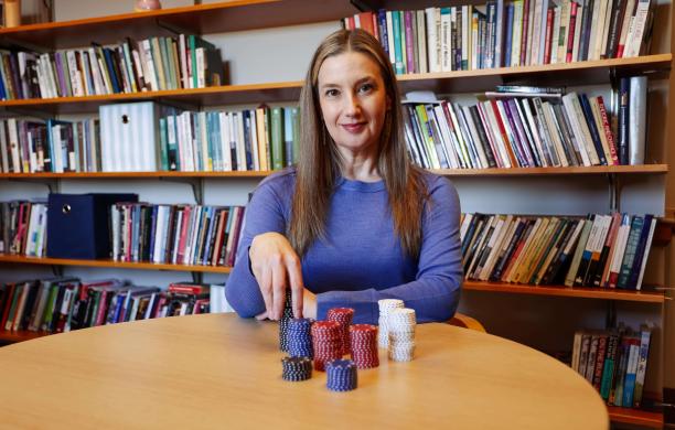 Brooke sits at a wooden table with poker chips in front of her and rests her hand on top of them; a bookshelf is behind her. 