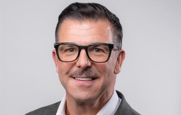 Jason wears black glasses, a dark, plaid suit jacket, and a crisp white button up. He smiles at the camera in a well lit room.