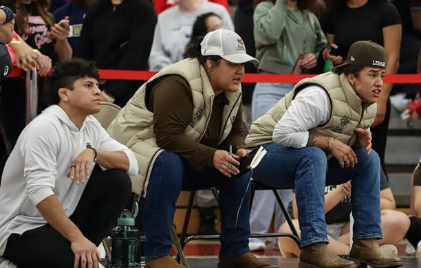 Three Lehigh wrestling coaches crouch at the edge of a gym mat, watching a match intently, while a crowd of spectators stands behind them.