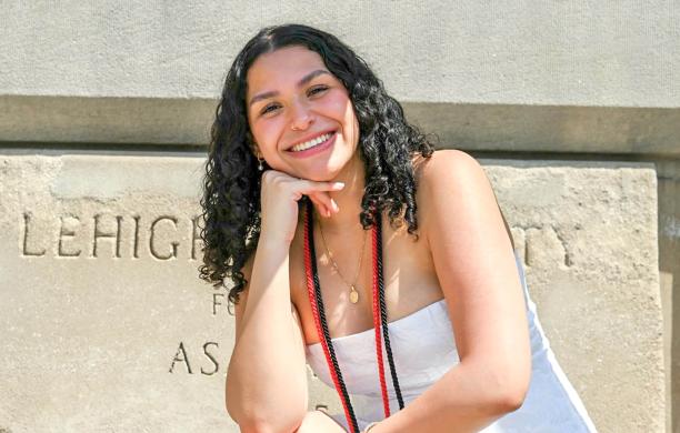 Arianna Morataya, wearing graduation cords around her neck, sits leaning her chin on her hand and smiling with the words “Lehigh University” seen behind her carved into concrete.