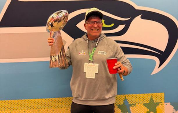 Todd Brunner holds a party cup, cigar, and Super Bowl trophy with the Seahawks logo in the background