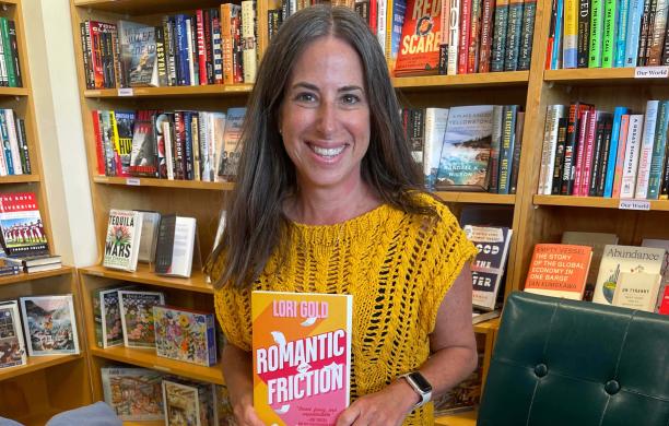 Lori wears a warm yellow, knit, short-sleeve sweater and holds up Romantic Friction. Behind her are lined bookshelves.