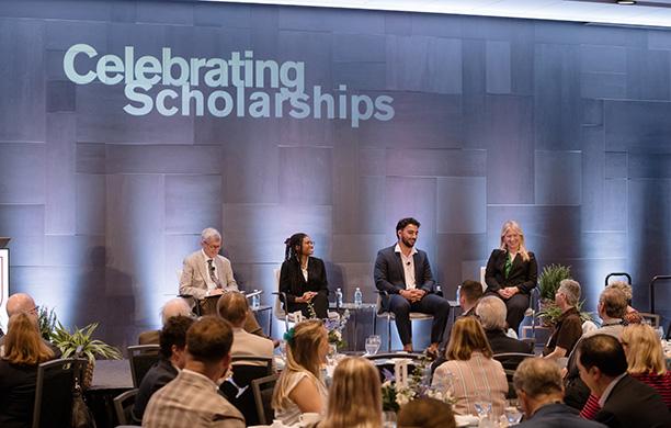 Four people sit on a stage in front of a crowd with the word Scholarship Celebration on the wall behind them
