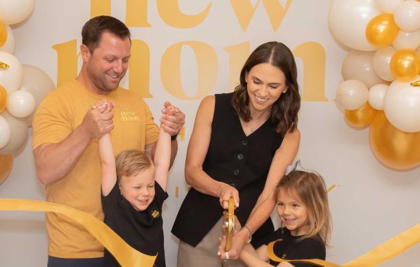Danielle with her husband and children cutting a yellow ribbon for the opening of New Mom School. They smile and wear merch. 