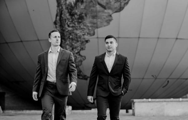 A black and white photo of Chris and Alfonso. They both wear business casual; dark blazers and slacks. Behind them is a globe. 