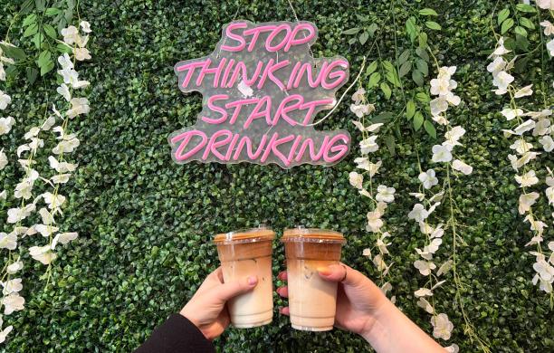 Two hands hold coffees in front of greenery. A sign that hangs above reads 'stop thinking, start drinking.' 