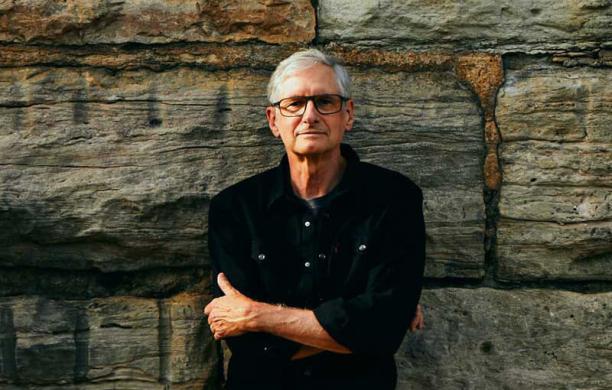 Jim Stanard wears a casual black button down shirt over a t-shirt while crossing his arms and leaning against a large stone wall.