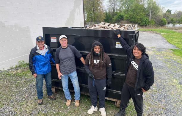 Jimmy and three other volunteers stand in front of a black dumpster. Two of the volunteers wear brown Lehigh shirts.