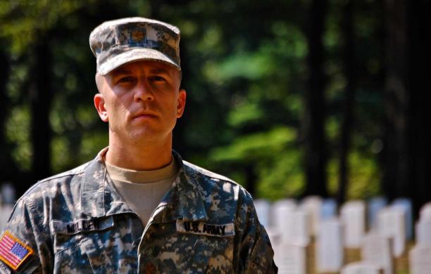 Nick wears his military fatigues in Arlington National Cemetery near headstones. His patrol cap casts his face in light shadow. 