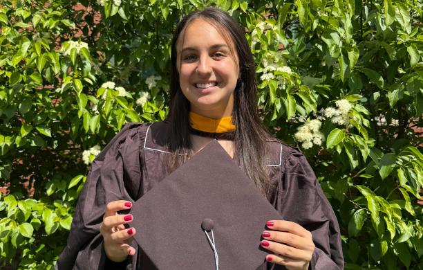 Gabby stands outside in her brown gown and holds the matching cap in her hands. Her nail polish is a bright plum.