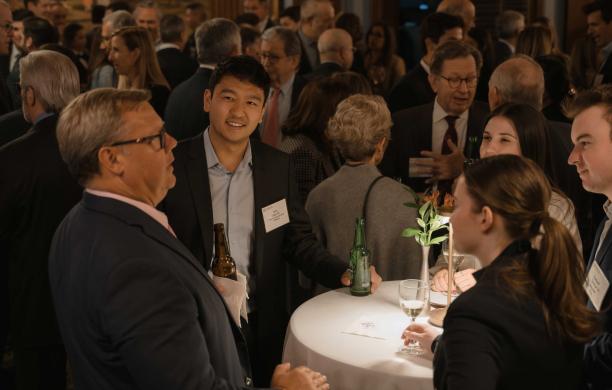 A group of alumni in a well-populated room. They all wear business casual and engage in conversation, some with drinks. 