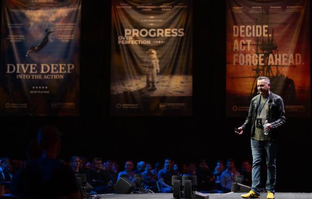 Holding a water bottle and wearing a lanyard, Joe Paluska stands on a dark stage with banners with inspirational mantras hanging behind him.