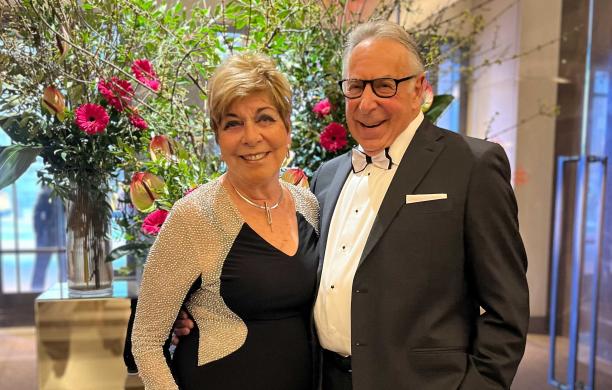 Michael wears a tuxedo with his arm around his wife Enid standing on his left side and dressed in a black and silver tight-fitting gown.