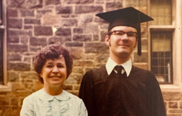 Holding his rolled diploma in his hand, Paul Elosge poses outdoors in graduation cap and gown next to a woman smiling with short dark hair in a powder blue dress. 