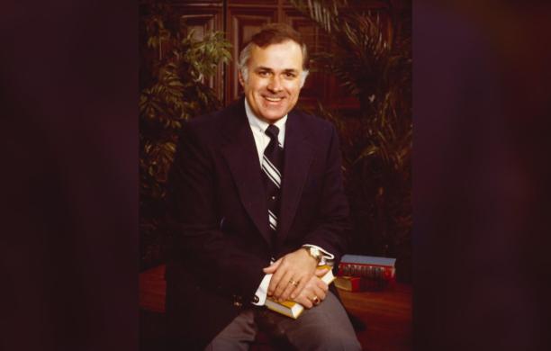 Posed on a stool with hands folded in his lap, holding a book, David Van Horn smiles for a faculty portrait in a sport coat and neck tie.
