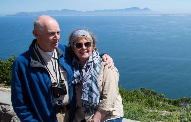 Jesse Salwen and wife Marty look off camera smiling while sitting together on a stone wall overlooking a body of water in Cape Town.
