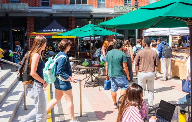 People walk around the Farmers' Market on a sunny day, many of them wearing backpacks. Bright green umbrellas shade tables.