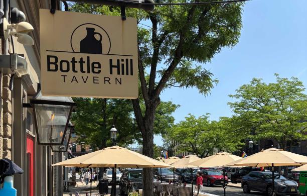 The outside of Bottle Hill Tavern on a sunny day. Their sign is visible alongside tables with umbrellas.