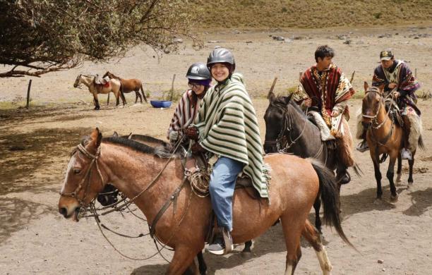 Lillian rides a horse in Ecuador while wearing a striped poncho and helmet. Other horses trail behind her.
