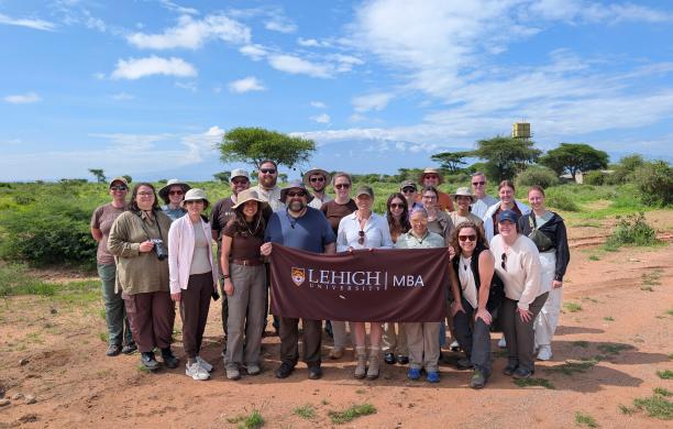A group of FLEX MBA students pose together outside in Kenya. They hold up a brown Lehigh MBA flag and smile. 