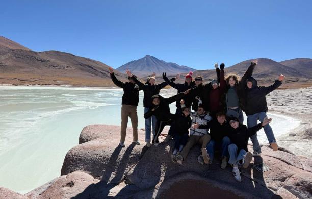 A group of students stand alongside a large blue lagoon in the mountains of Chile. They wave their hands in the air and smile.