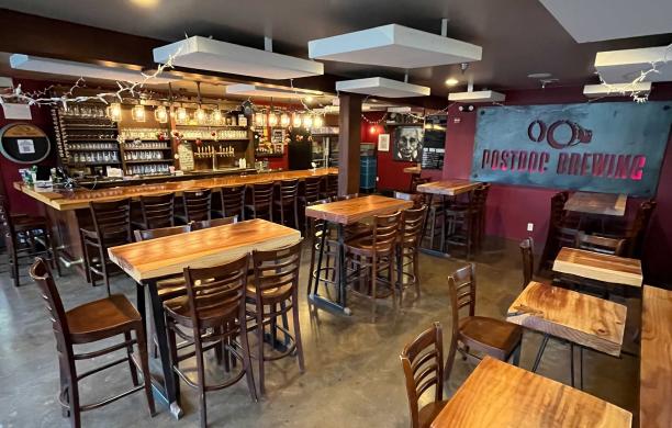 The interior of Postdoc Brewing features warm lighting, wood tables and chairs, and a fully stocked bar with high top seating.